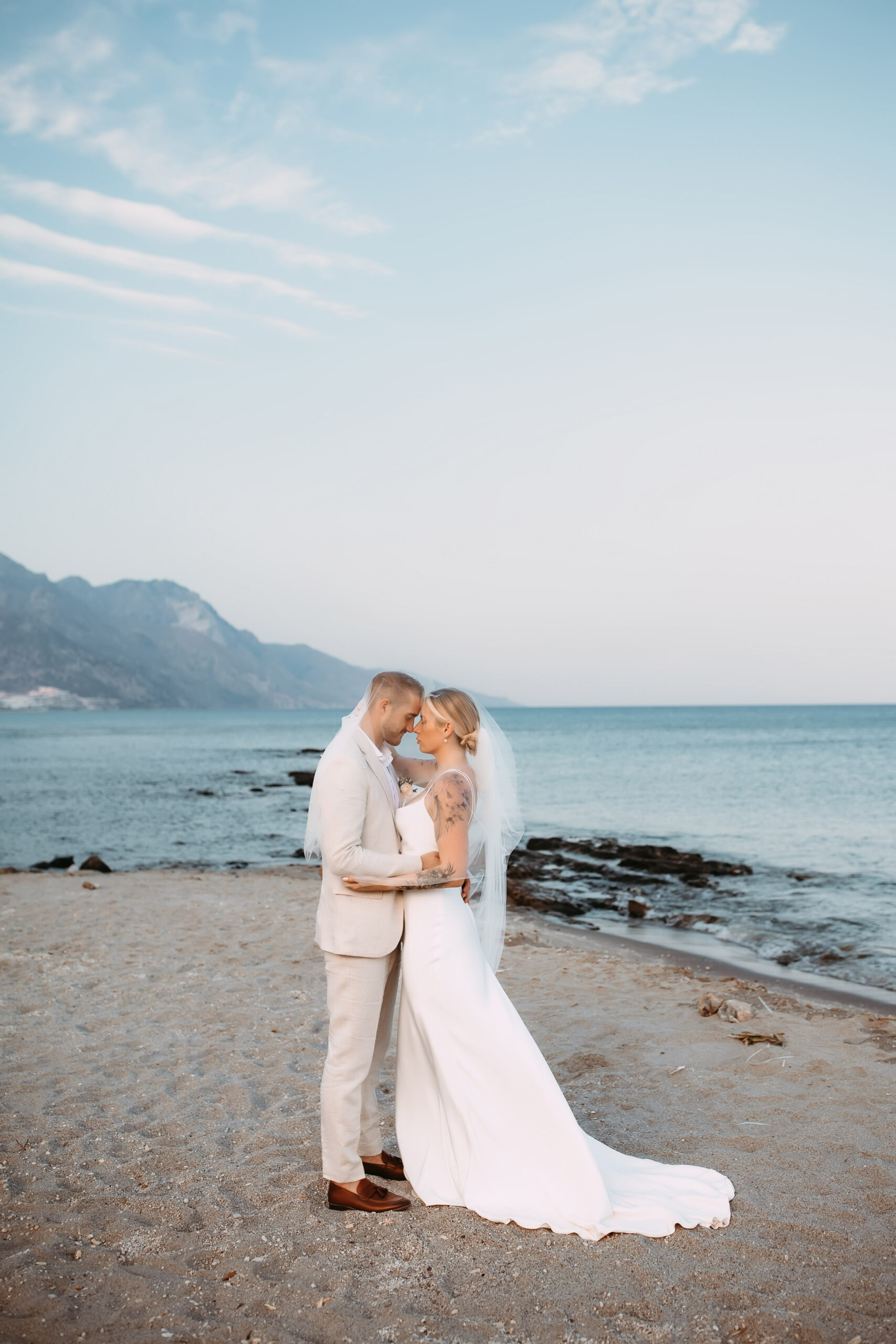 Bride and groom embracing on the beach at sunset after their destination wedding in Kos, Greece, with the sea and mountains in the background.